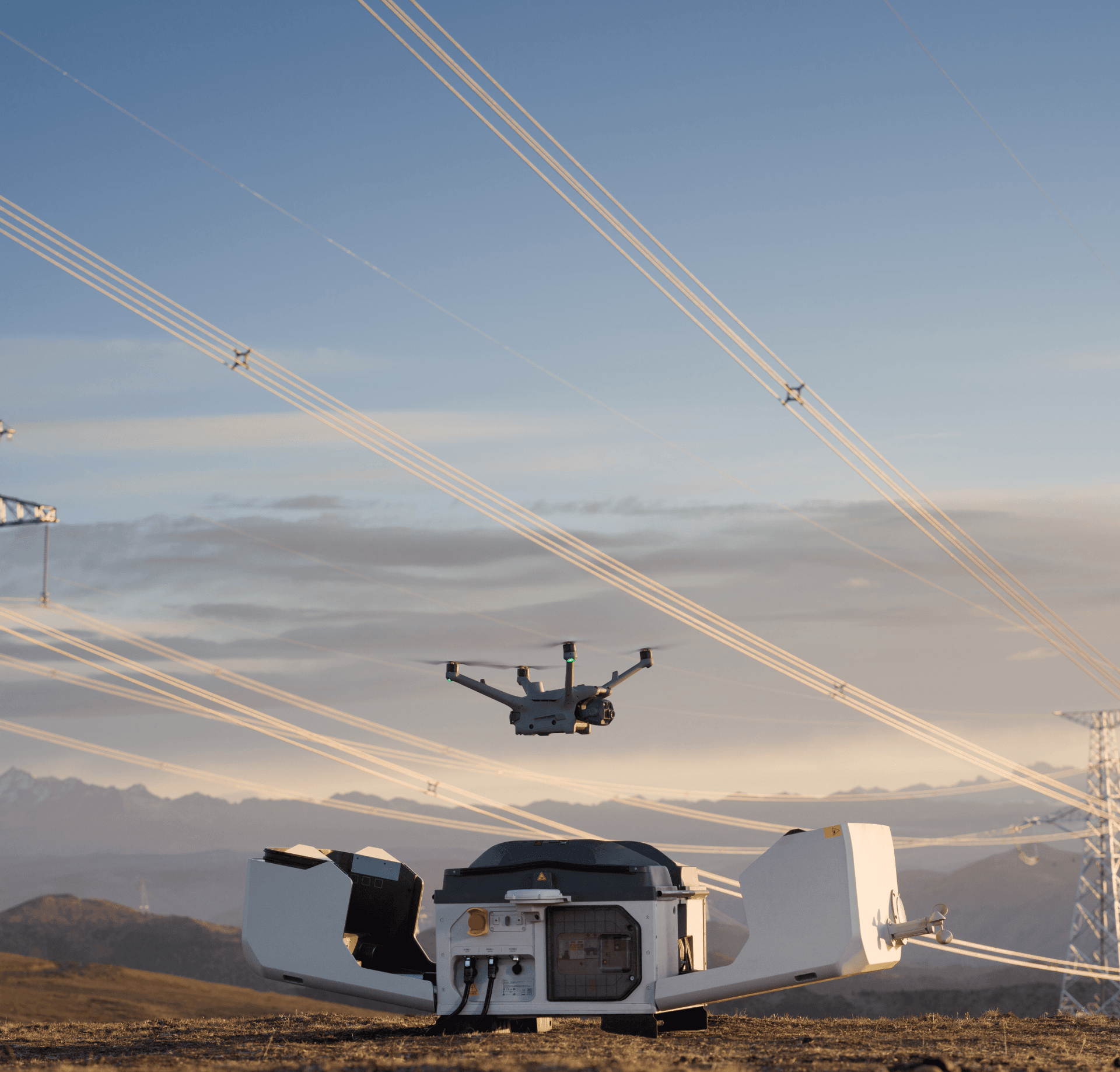 En drönare lyfter från en landningsstation i ett öppet landskap med kraftledningar i bakgrunden.