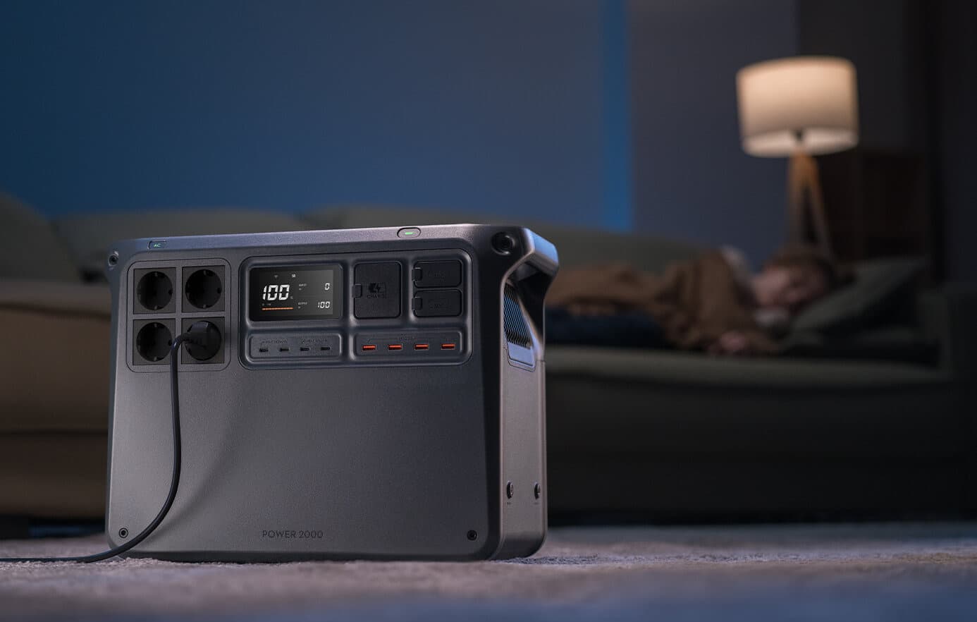 En kraftfull bärbar DJI Power 2000 står på golvet i ett dämpat upplyst rum, medan en person sover på en soffa i bakgrunden.