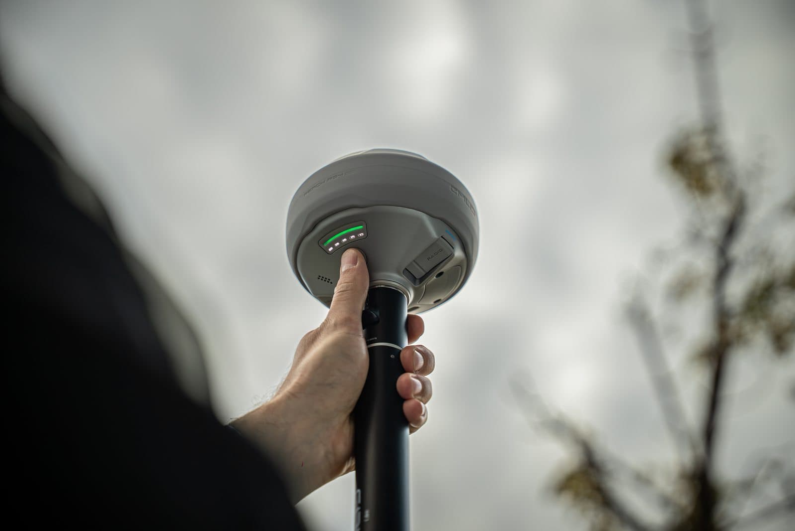 En hand håller i en GPS-mottagare mot en molnig himmel, med indikatorlampor som lyser grönt.