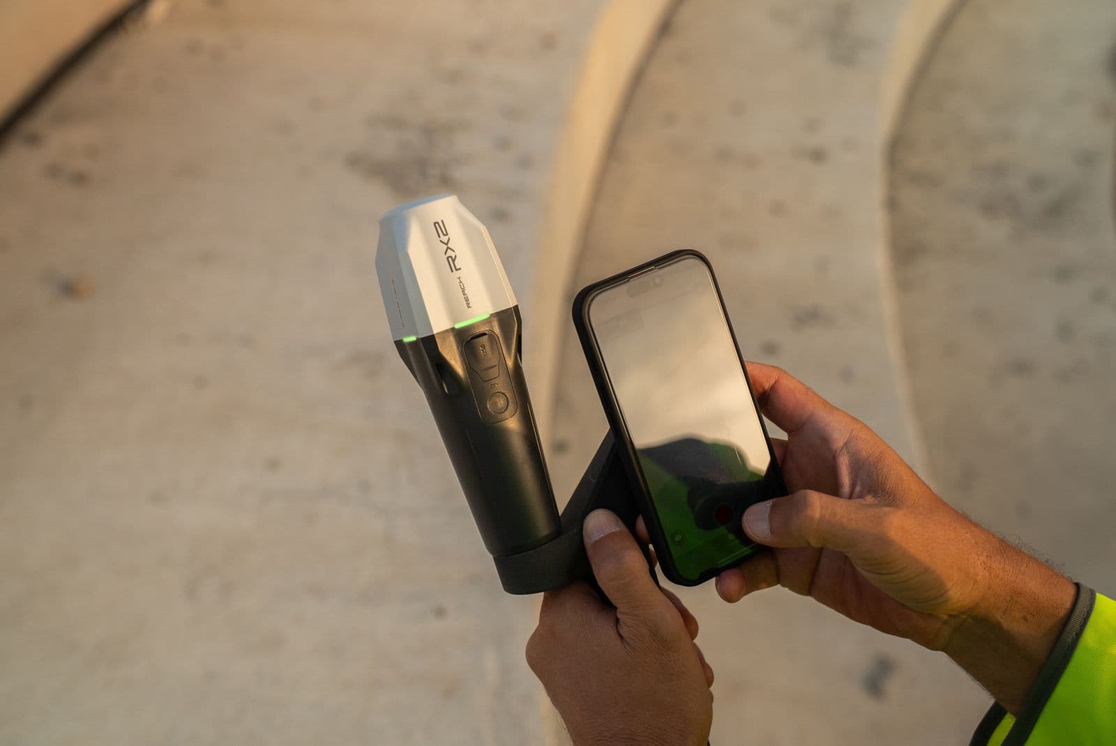 En person håller en smartphone och en handhållen Emlid Reach RX2 -enhet med texten 'RX2' i en utomhusmiljö.