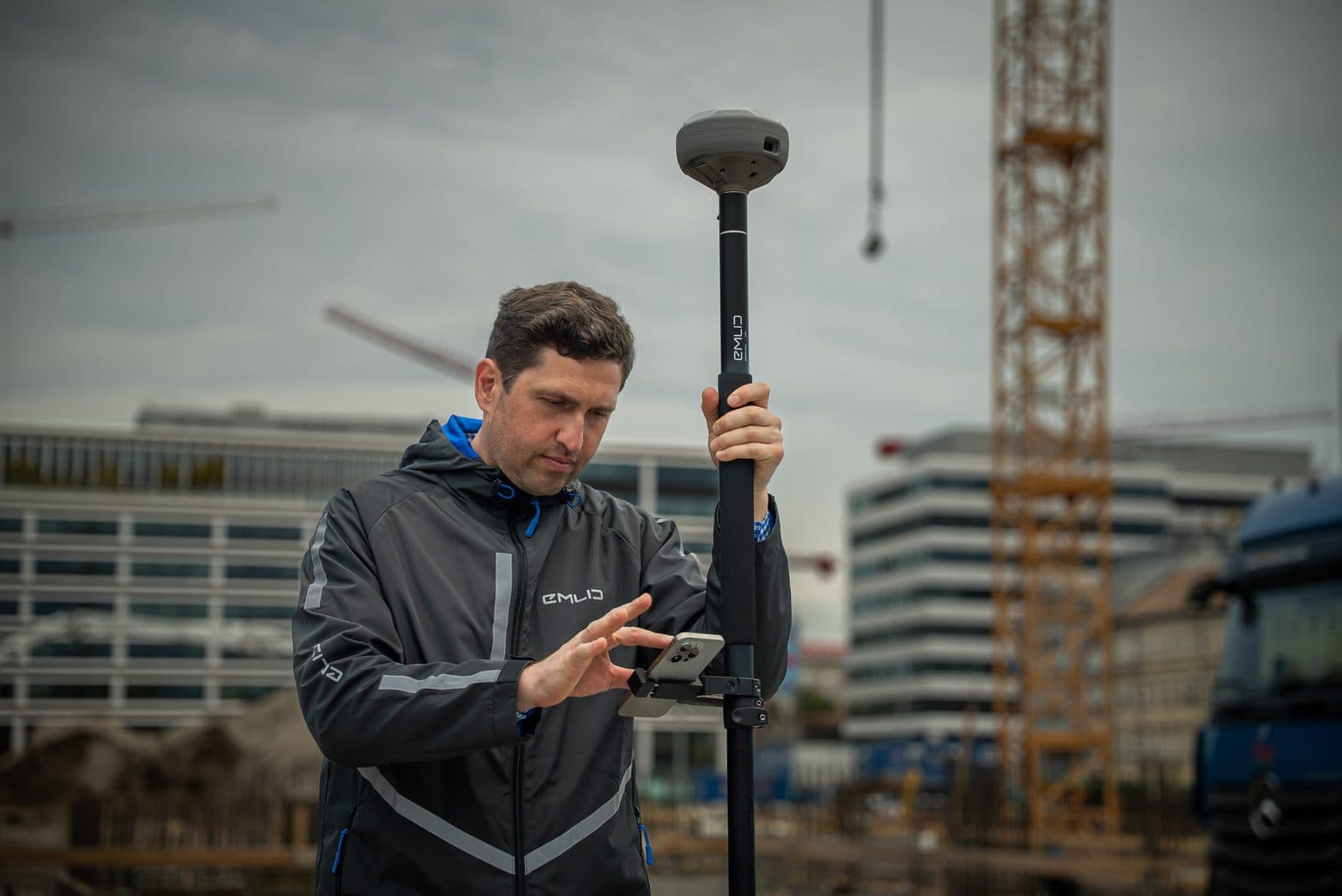 En person använder en GPS-mottagare på en byggarbetsplats, omgiven av kranar och moderna byggnader.