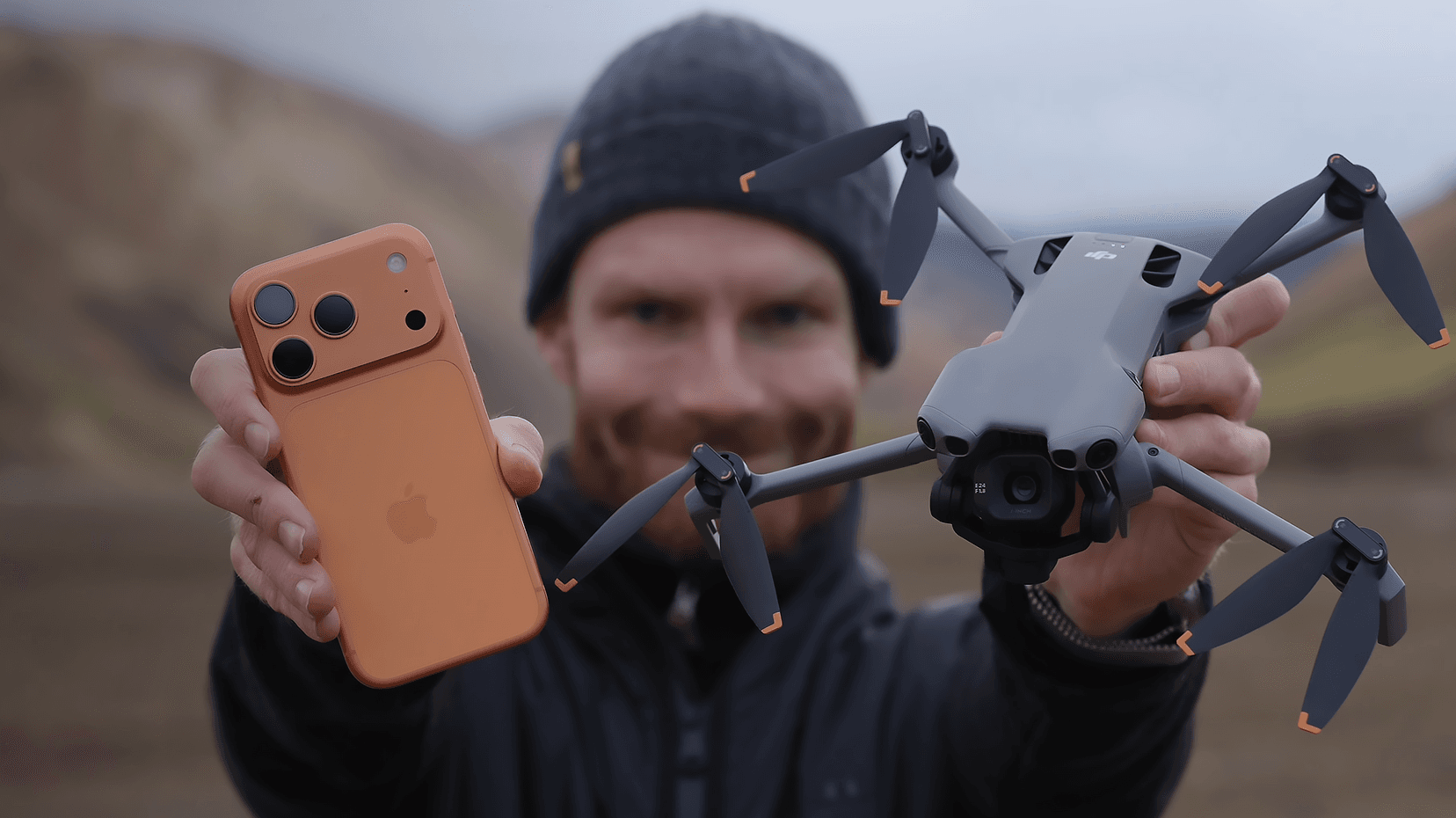 En person håller upp en orange smartphone och en grå drönare i en utomhusmiljö.