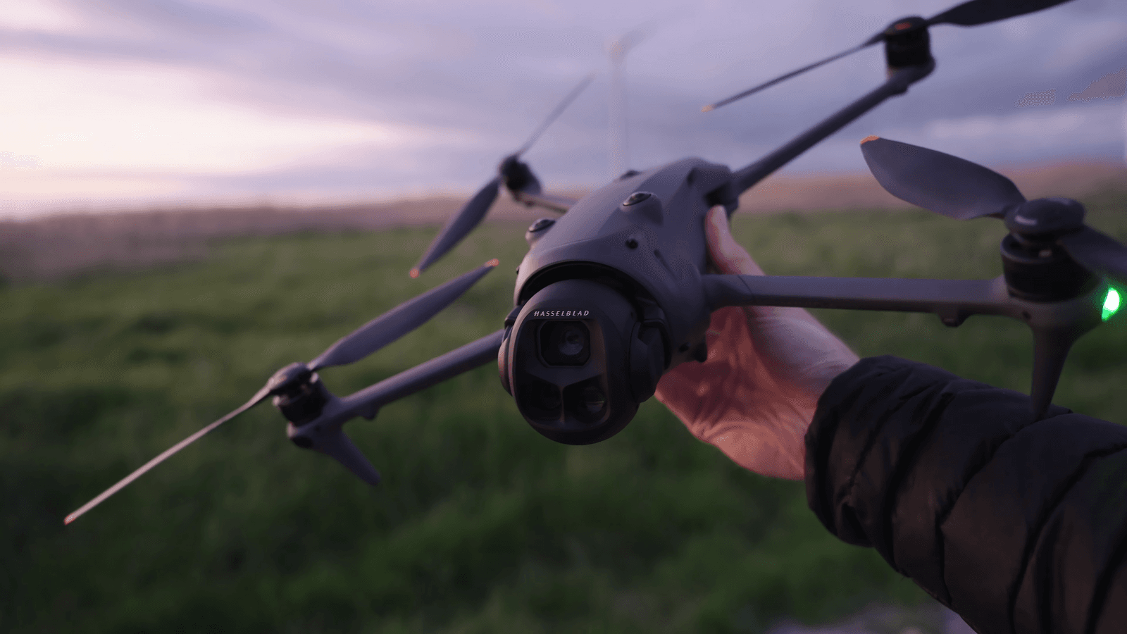 En person håller en DJI Mavic 4 Pro-drönare med en kamera i ett öppet landskap vid solnedgång.