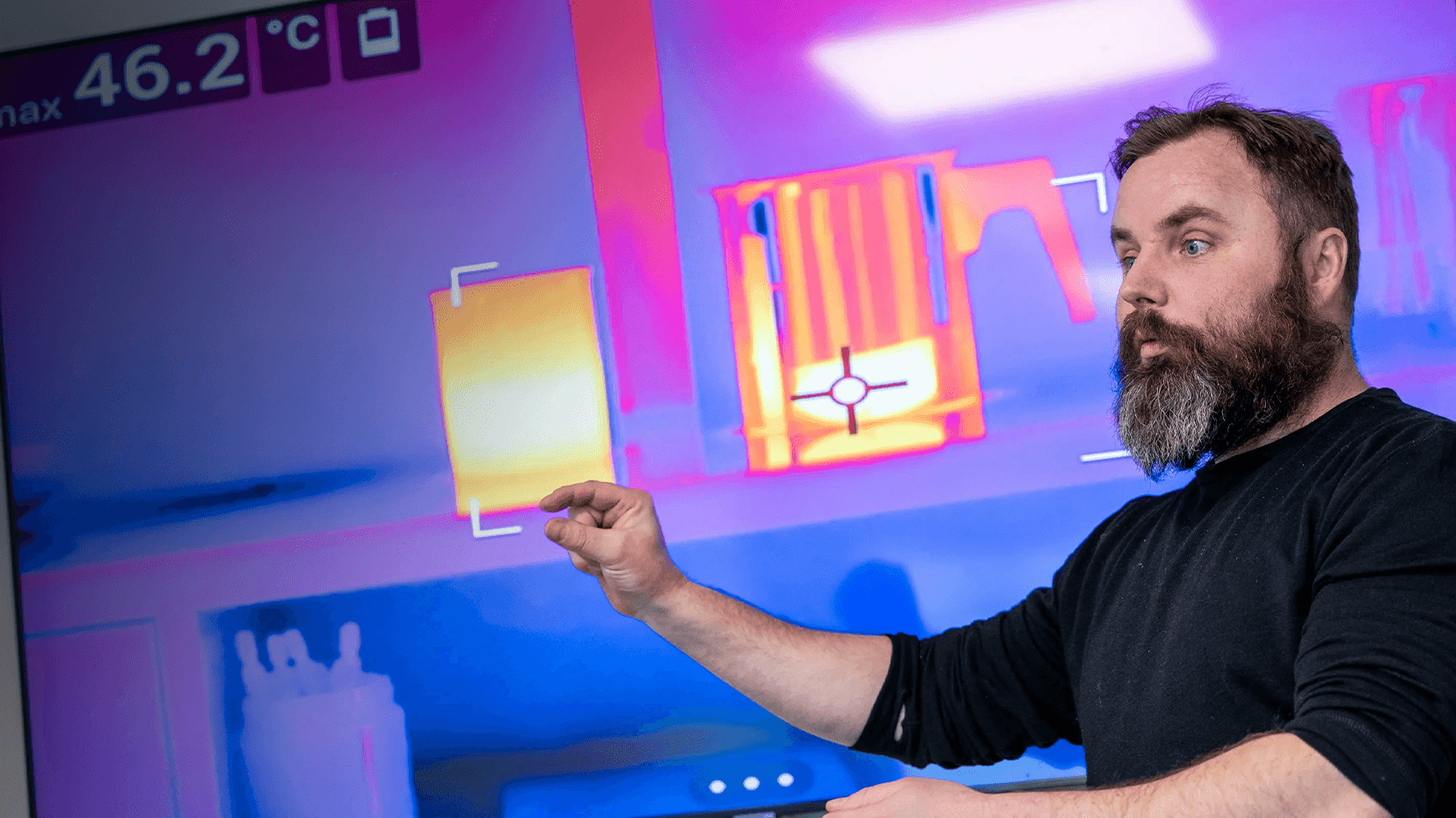 A person gestures towards a thermal imaging display showing temperature variations, with a maximum reading of 46.2°C.